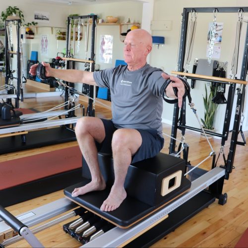 Jim sitting on the reformer with hands in the straps with arms out wide